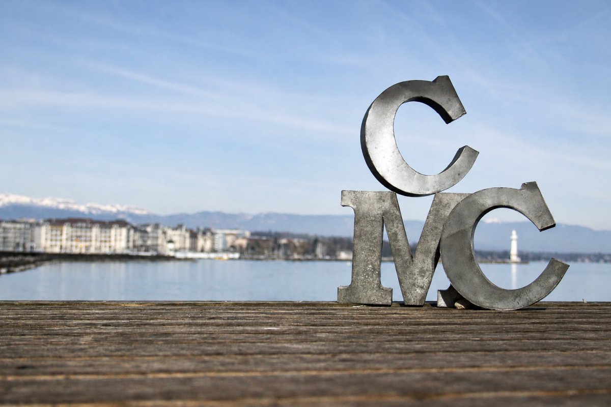 les lettres cmc, posées sur un ponton de la rive gauche du lac Léman, encadrent le phare des Pâquis au loin.